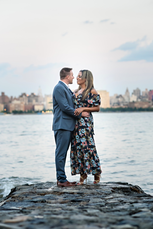 A man holds his bride to be on the rocks along the hudson river with NYC in the background.